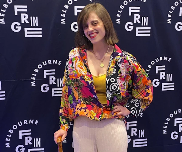 Liel Bridgford has shoulder length brown hair and wears a colourful shirt while posing in front of a Melbourne Fringe Festival banner. She holds a walking cane and smiles at the camera.