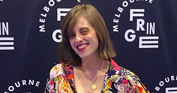 Liel Bridgford has shoulder length brown hair and wears a colourful shirt while posing in front of a Melbourne Fringe Festival banner. She holds a walking cane and smiles at the camera.