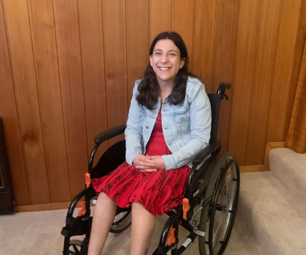 Laura Petenuzzo, a disability advocate and writer, sits smiling in her wheelchair. She has long dark brown hair and wears a red dress with a denim jacket.