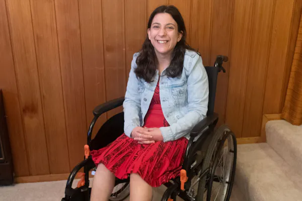 Laura Petenuzzo, a disability advocate and writer, sits smiling in her wheelchair. She has long dark brown hair and wears a red dress with a denim jacket.