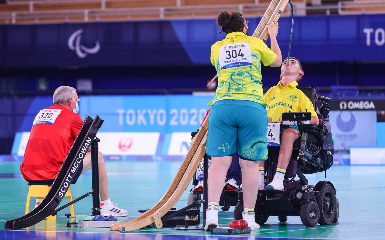 Daniel has short hair and is wearing the Australian Paralympic green and yellow uniform. He is holding a stick in his mouth to push a boccia ball down a wooden ramp. His ramp assistant Ashlee is holding the ramp upright. She has dark hair and is wearing the same outfit as Daniel. Daniel is a wheelchair user.