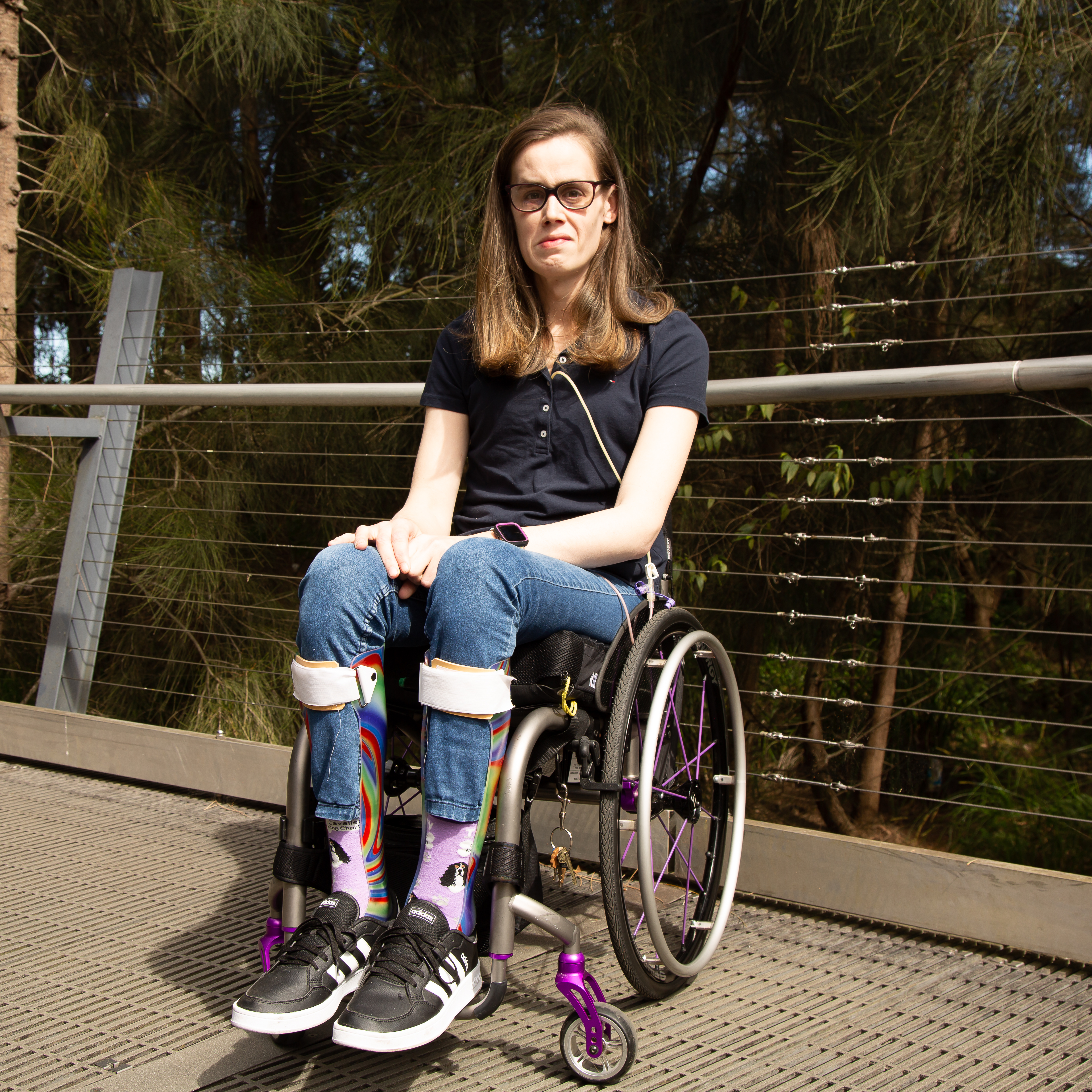 A woman, Kim, sits in a wheelchair and looks at the camera. She's wearing a t shirt and jeans with colourful socks. 