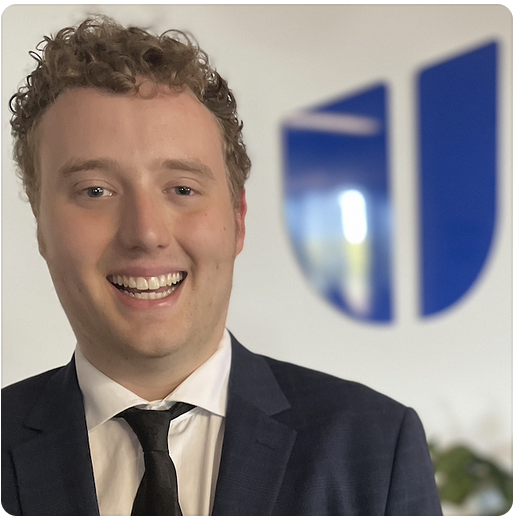 Julian is smiling, standing in front of the blue Hireup logo. He has short, curly blonde hair and is wearing a navy suit and black tie.