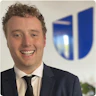 Julian is smiling, standing in front of the blue Hireup logo. He has short, curly blonde hair and is wearing a navy suit and black tie.