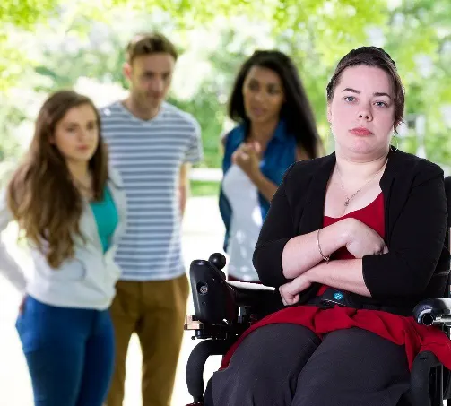 A group of people are pointing at another person using a wheelchair. The person in the wheelchair has her arms folded and looks sad.