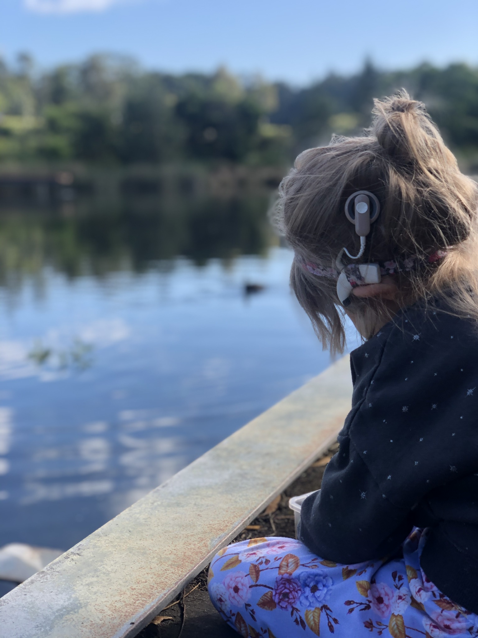 A young child is sitting by a pond. They're wearing a jumper and have a cochlear implant.