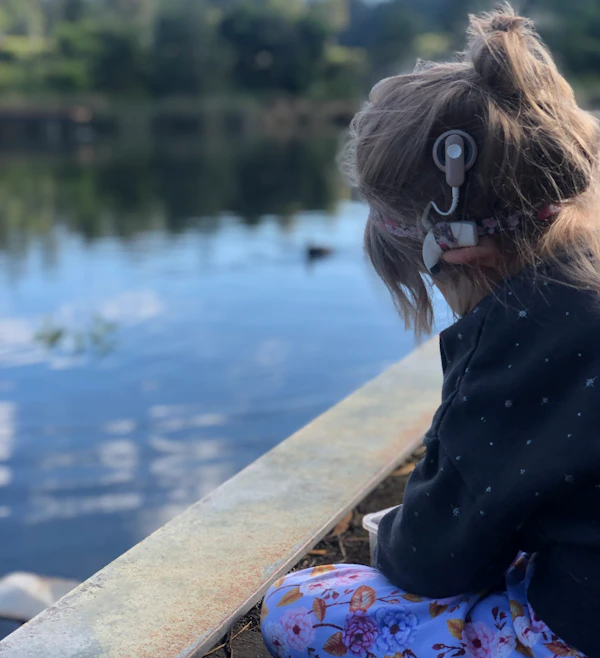 A young child is sitting by a pond. They're wearing a jumper and have a cochlear implant.