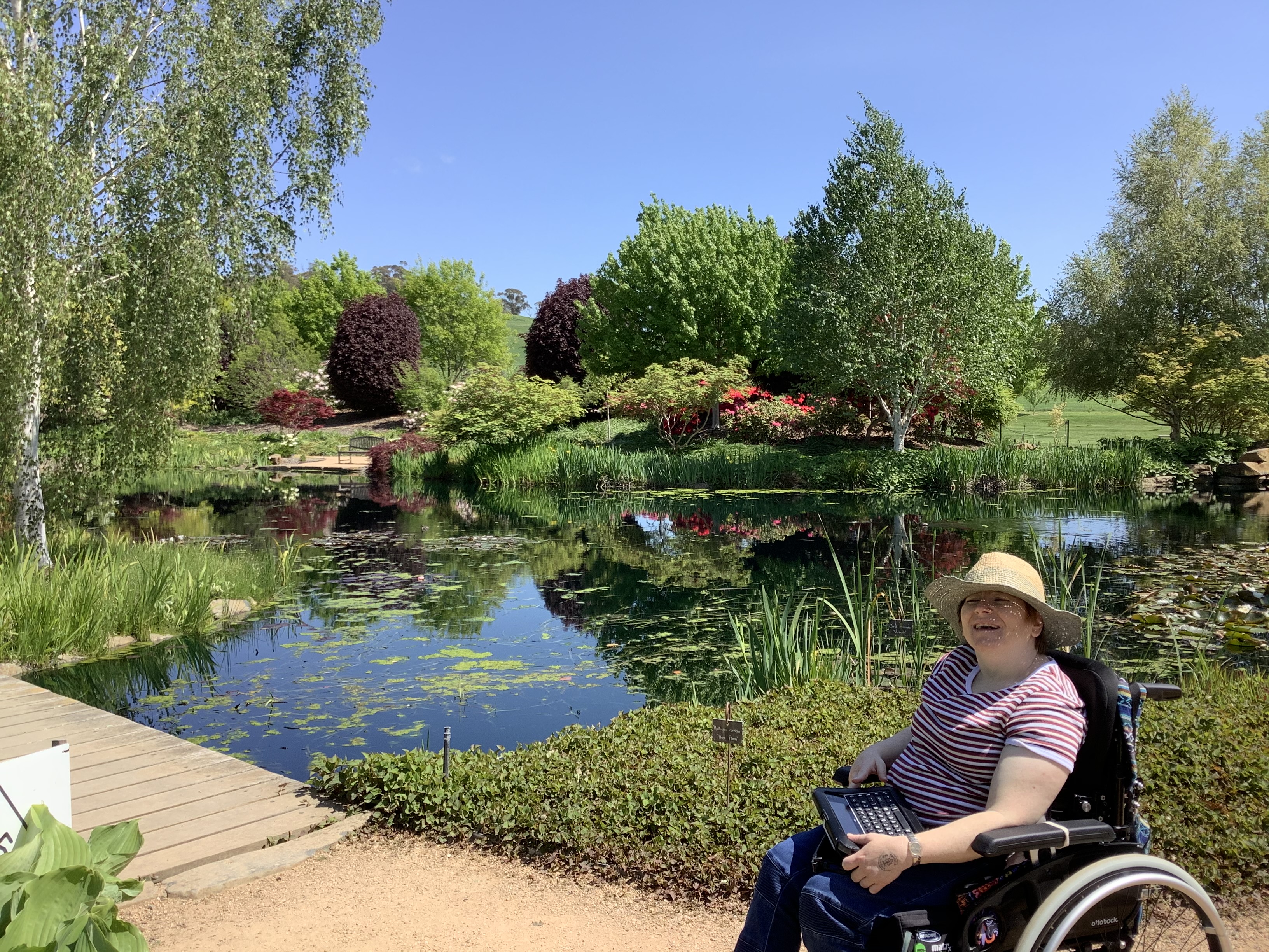 Fiona is wearing a candy coloured striped t-shirt and straw sun hat. She is smiling widely at the camera. Behind her is a pond full of lily pads and a large garden full of greenery. Fiona is a wheelchair user and has a speech keyboard resting on her lap. 