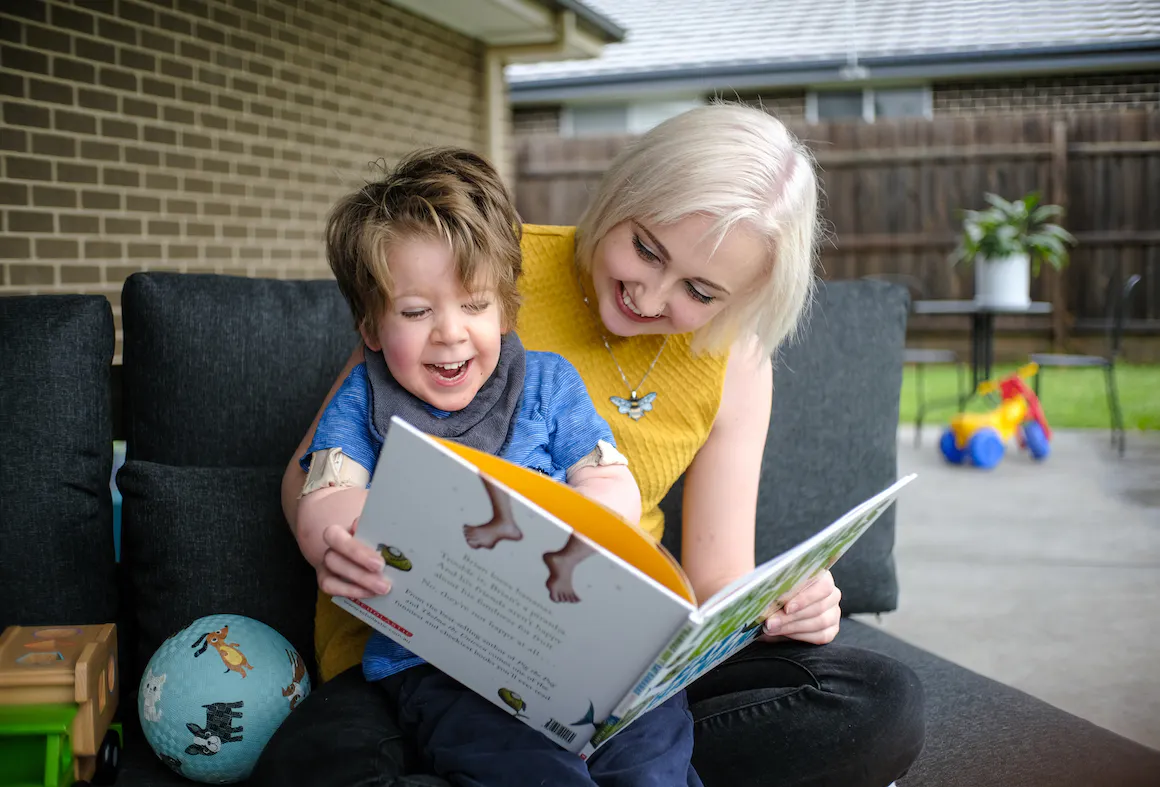 Arlo is wearing a light coloured top and dark pants. He's laughing and is reading a book that's held by his support worker Maddy. Maddy is also laughing. 