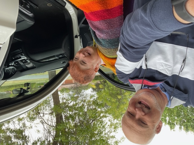 Brian and Jill are smiling getting into a white car. Brian has a shaved head and is wearing a blue gray and white striped hoodie. Jill has short orange hair and is wearing a rainbow striped jumper.