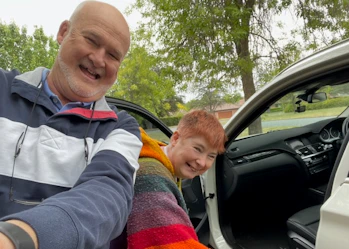 Brian and Jill are smiling getting into a white car. Brian has a shaved head and is wearing a blue gray and white striped hoodie. Jill has short orange hair and is wearing a rainbow striped jumper.