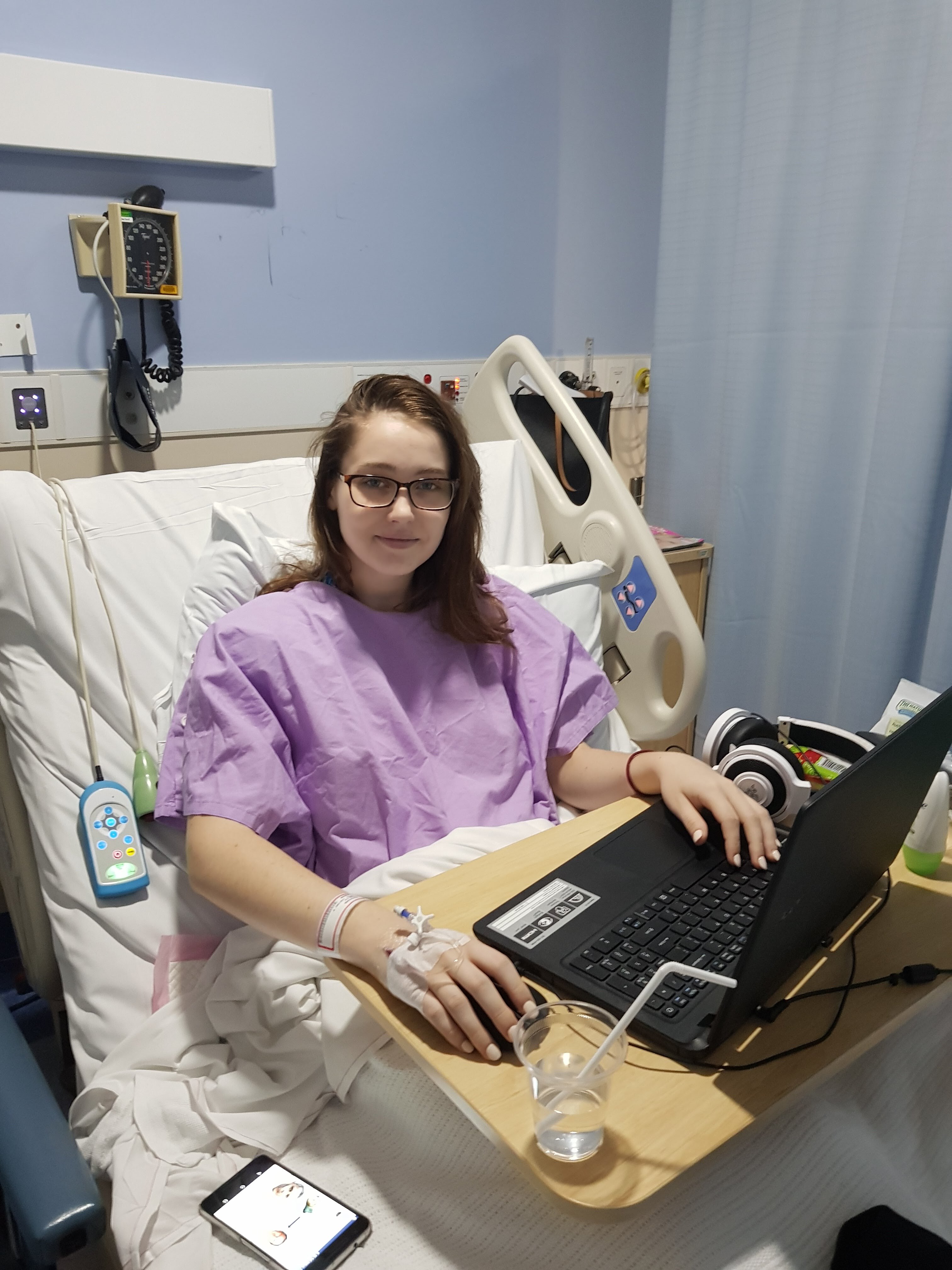 Megan sits in a hospital bed on her laptop. She is looking towards the camera. 