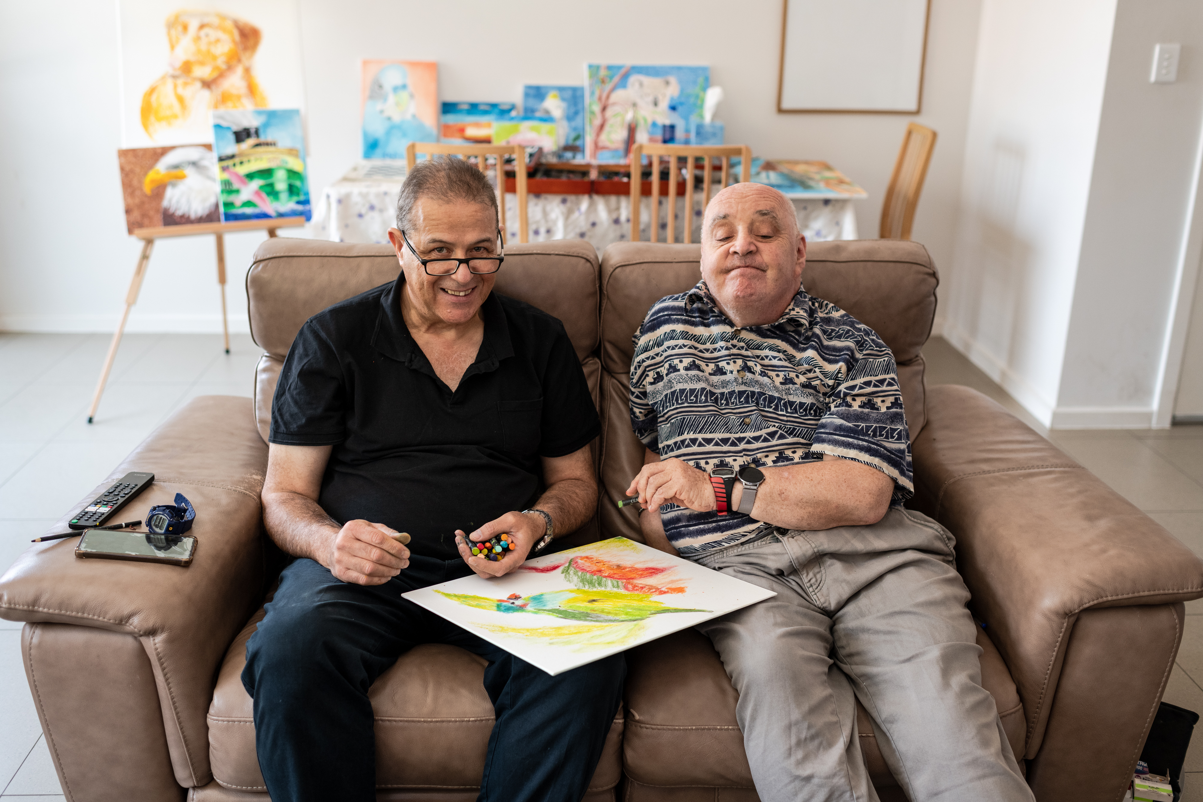 Daniel and Graeme are sitting on a brown couch doing art together with oil pastels. Daniel has short brown hair, wears reading glasses, a black collared shirt and navy trousers. Graeme has short white hair and is wearing two digital watches, a blue and white patterned shirt and grey trousers. 