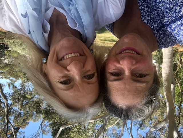 Justine is wearing lipstick plus a white top with blue flowers. Narelle has a blue scarf and earrings to match. Both women are smiling and have their heads close together for the selfie. In the background is a lush garden.