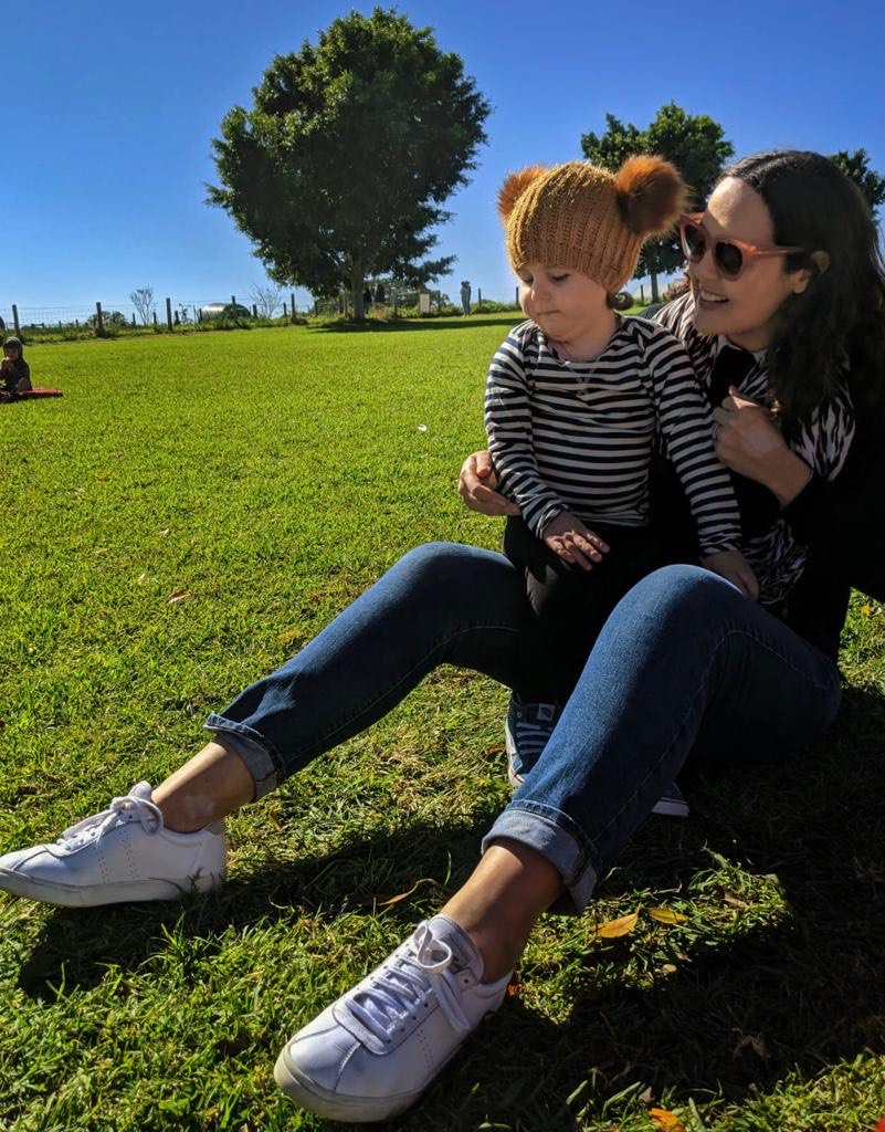 Chelsey is wearing sunglasses and white joggers. Coco is resting on Chelsea's knee and wears a beanie with two pom poms. They're on the grass in a green field.