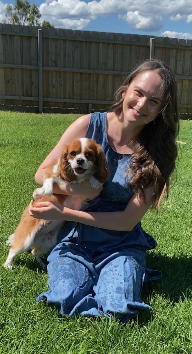 Tayla is holding a light brown and white cocker spaniel. She is kneeling on a green lawn and is wearing a blue jumpsuit. In the background is a tall wooden fence.