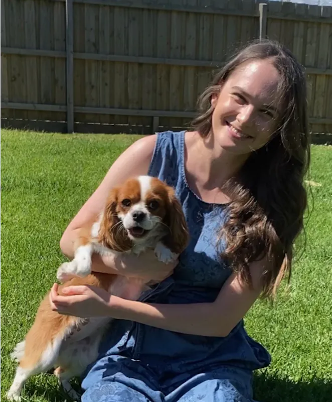 Tayla is holding a light brown and white cocker spaniel. She is kneeling on a green lawn and is wearing a blue jumpsuit. In the background is a tall wooden fence.