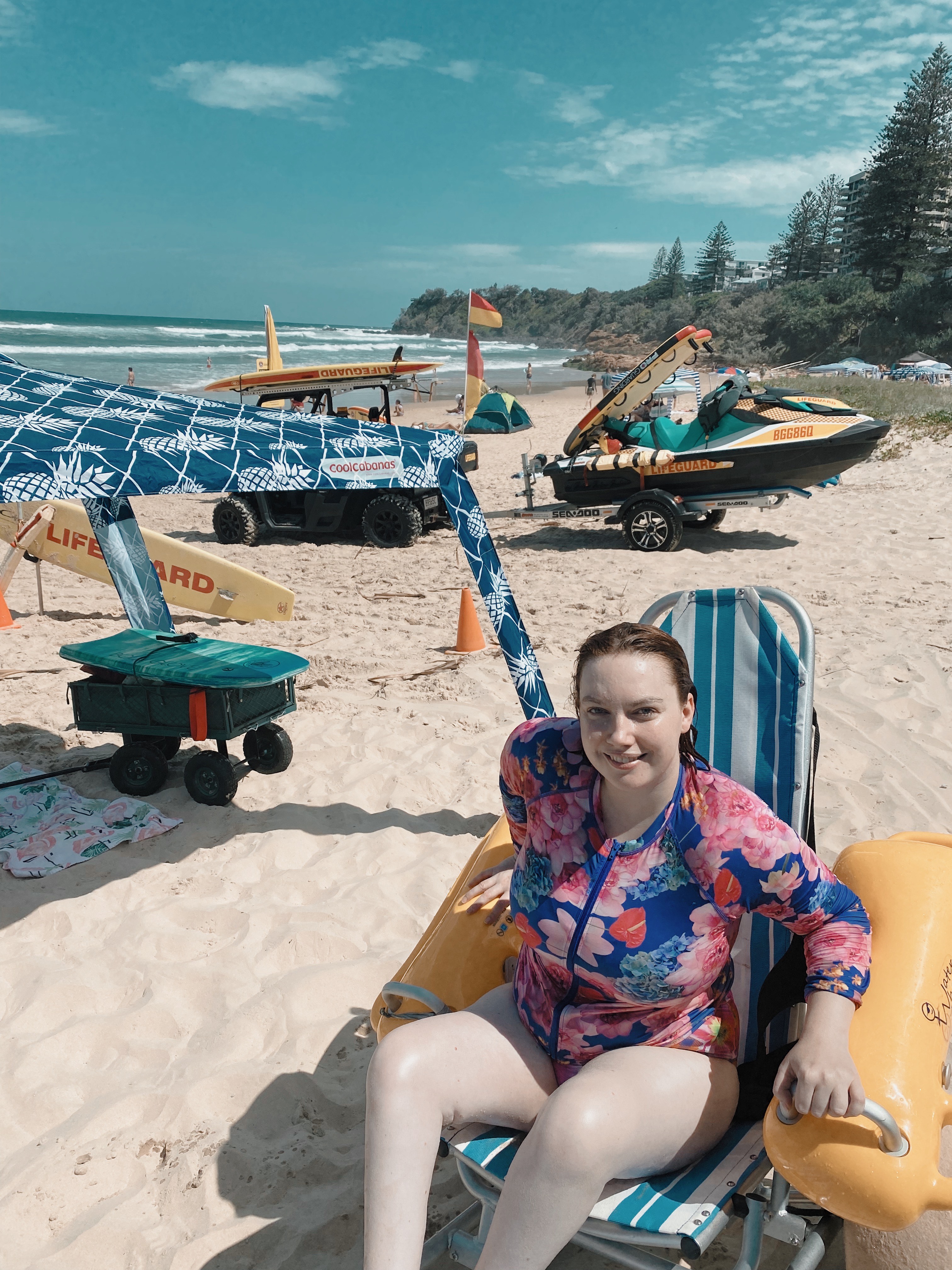 Elly Desmarchelier sits in a beach wheelchair on the sand with the ocean behind her. She wears a floral swimsuit and smiles at the camera. 