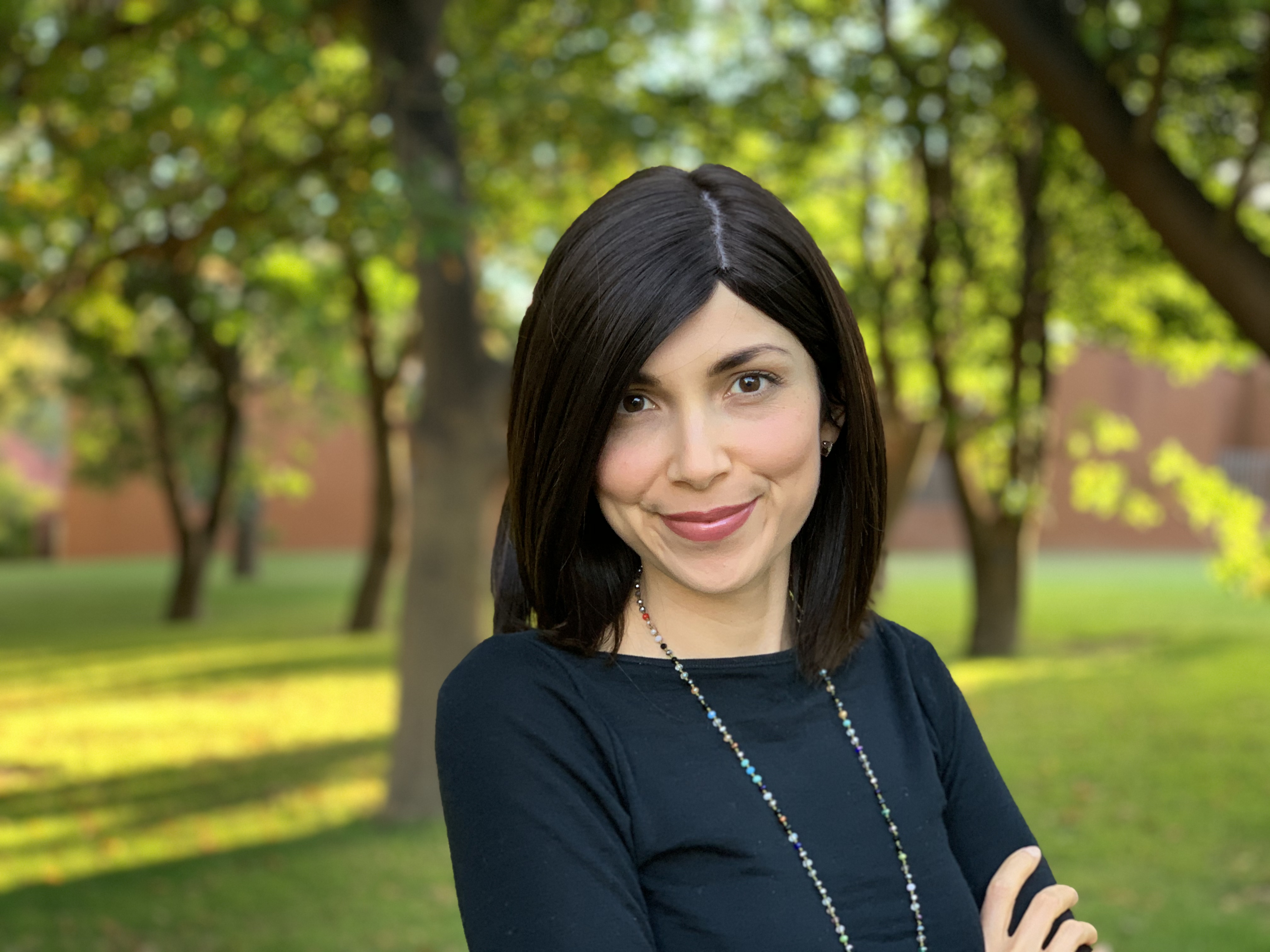 Natalia has short brown hair and is wearing a multi-coloured beaded necklace. Natalia is standing in the shade of trees. 