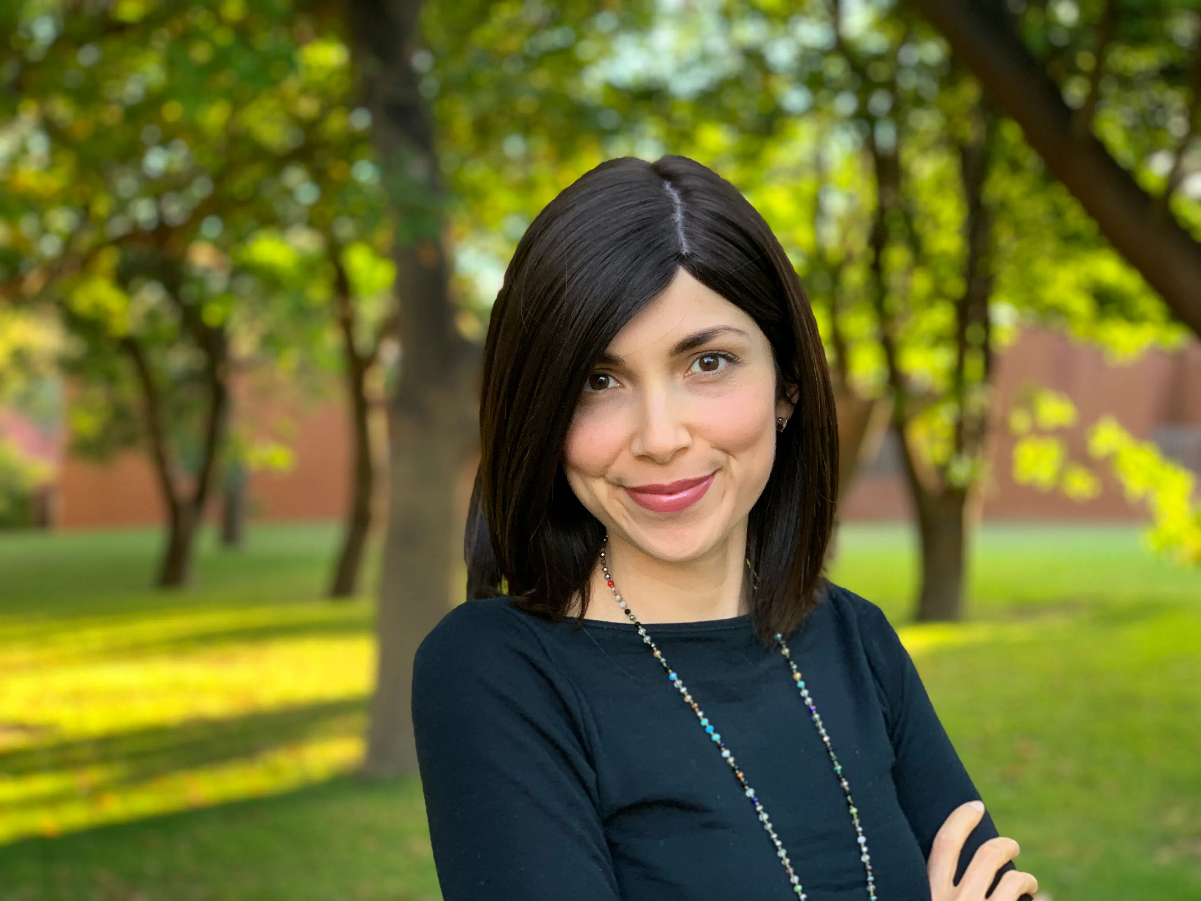 Natalia has short brown hair and is wearing a multi-coloured beaded necklace. Natalia is standing in the shade of trees.