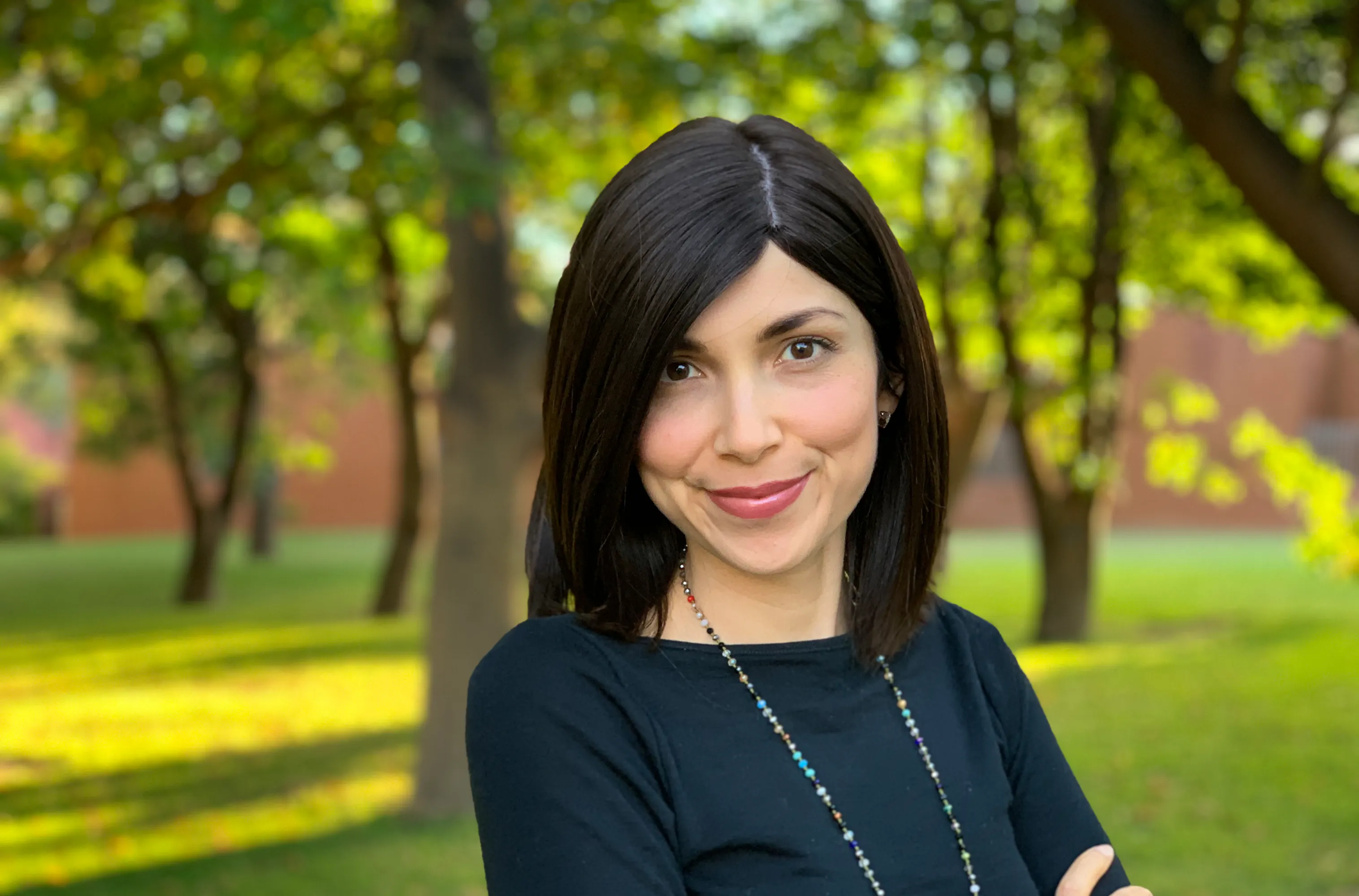 Natalia has short brown hair and is wearing a multi-coloured beaded necklace. Natalia is standing in the shade of trees.