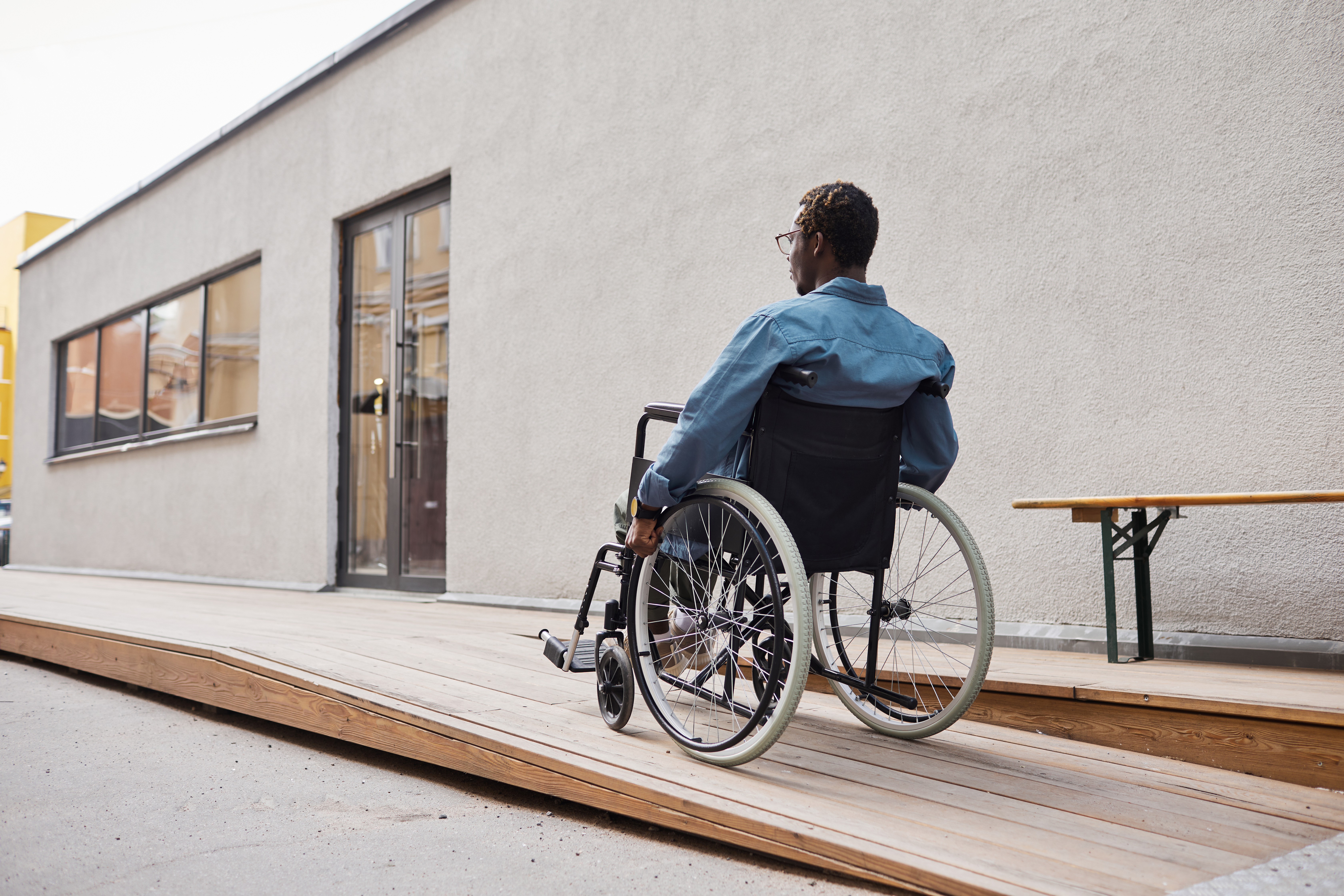 A man in a blue collared shirt is moving up a wooden ramp towards a door. The man is a wheelchair user and is wearing glasses. The building behind has grey cement walls.