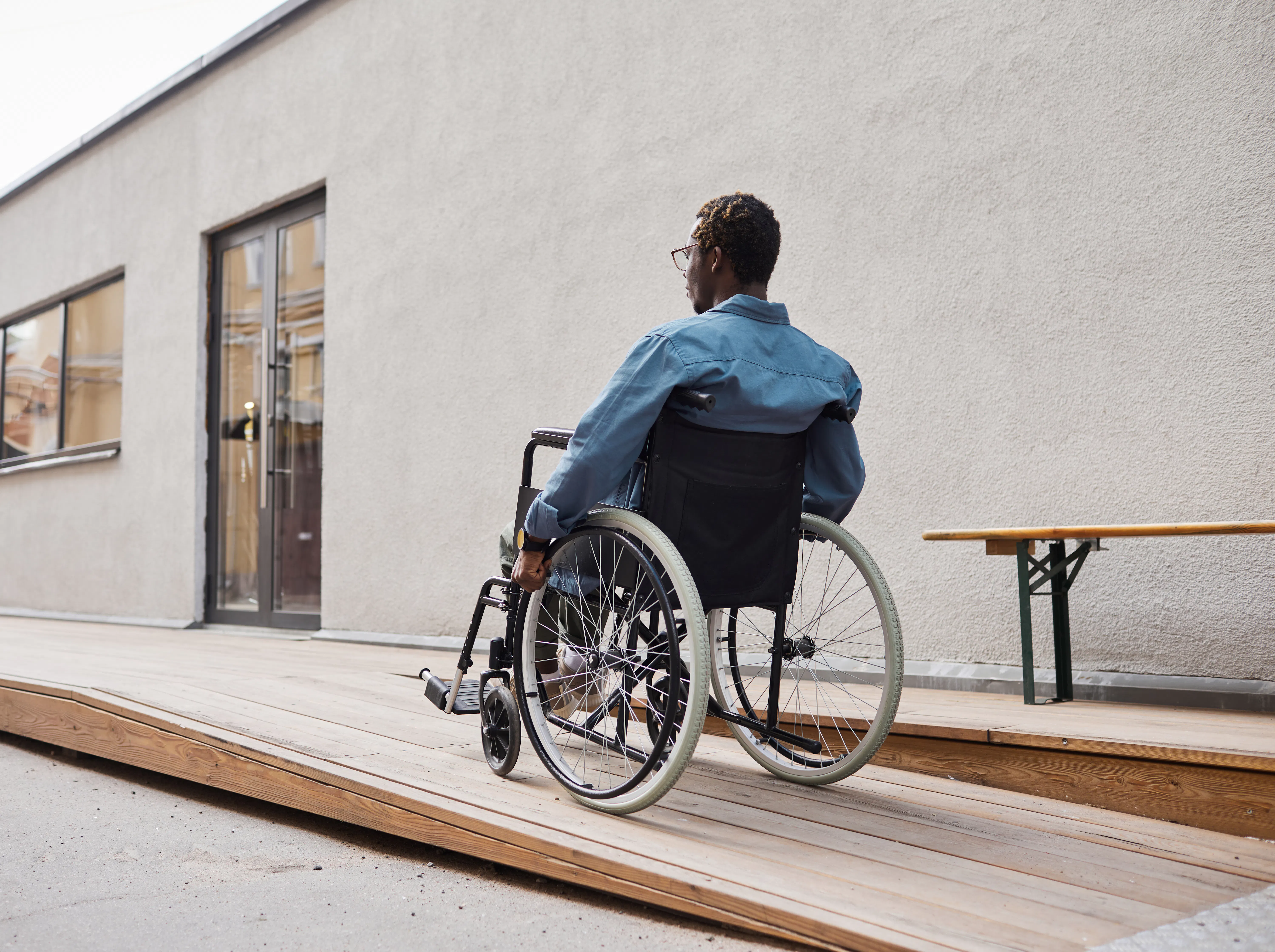 A man in a blue collared shirt is moving up a wooden ramp towards a door. The man is a wheelchair user and is wearing glasses. The building behind has grey cement walls.