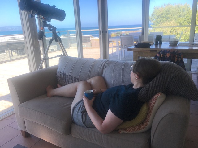 A woman lies on a couch looking out at an ocean. She is holding a mug and there's a telescope at the end of the couch. 