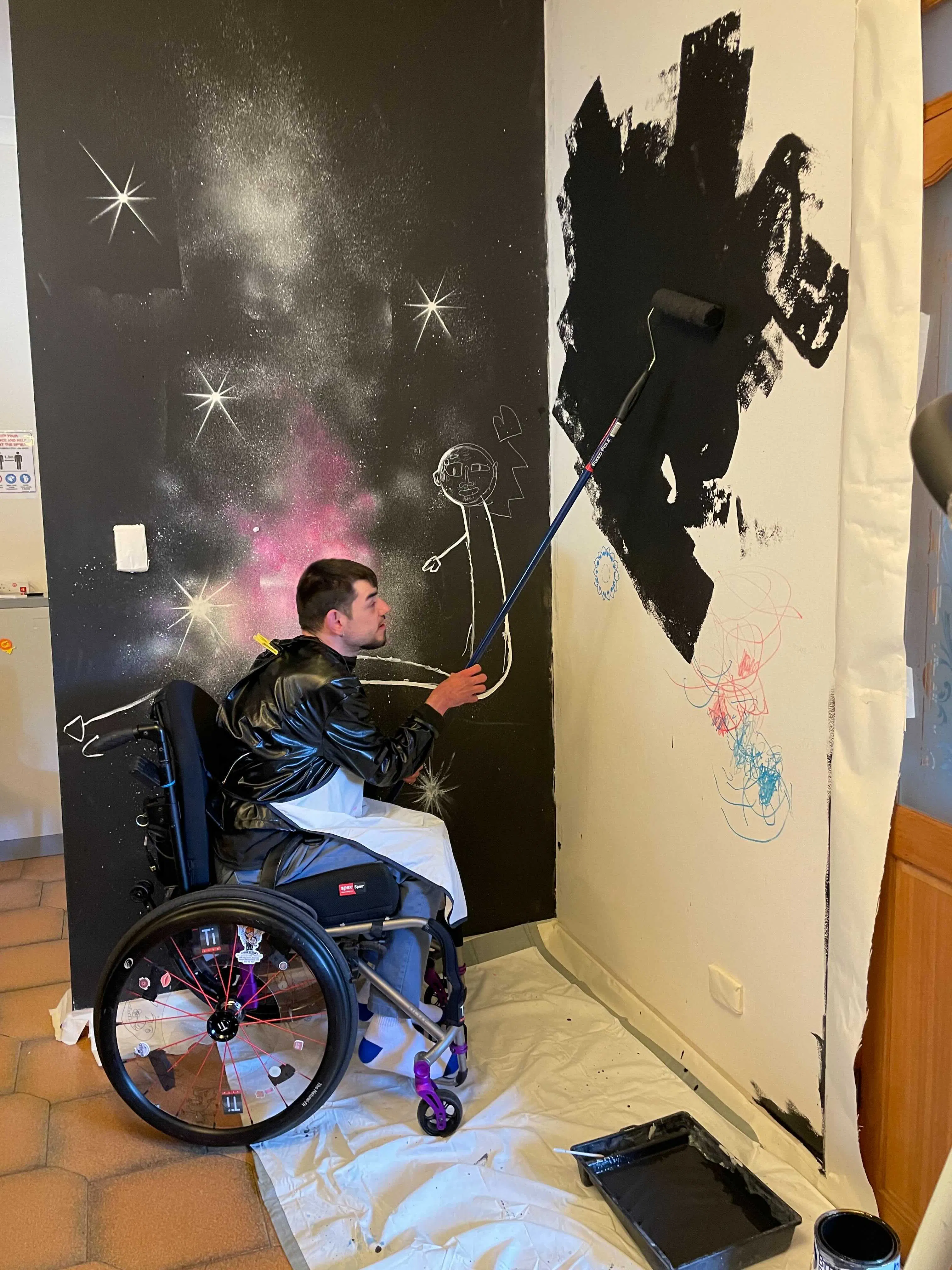 Andy is applying the black base coat of the mural with a paint roller. There is a sheet protecting the tile floor
