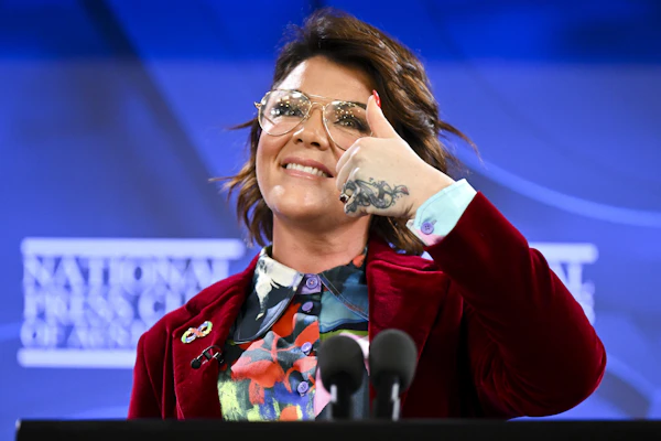 Image Shows: Em Rusciano standing at the podium at the National Press Club, smiling and giving a thumbs up.