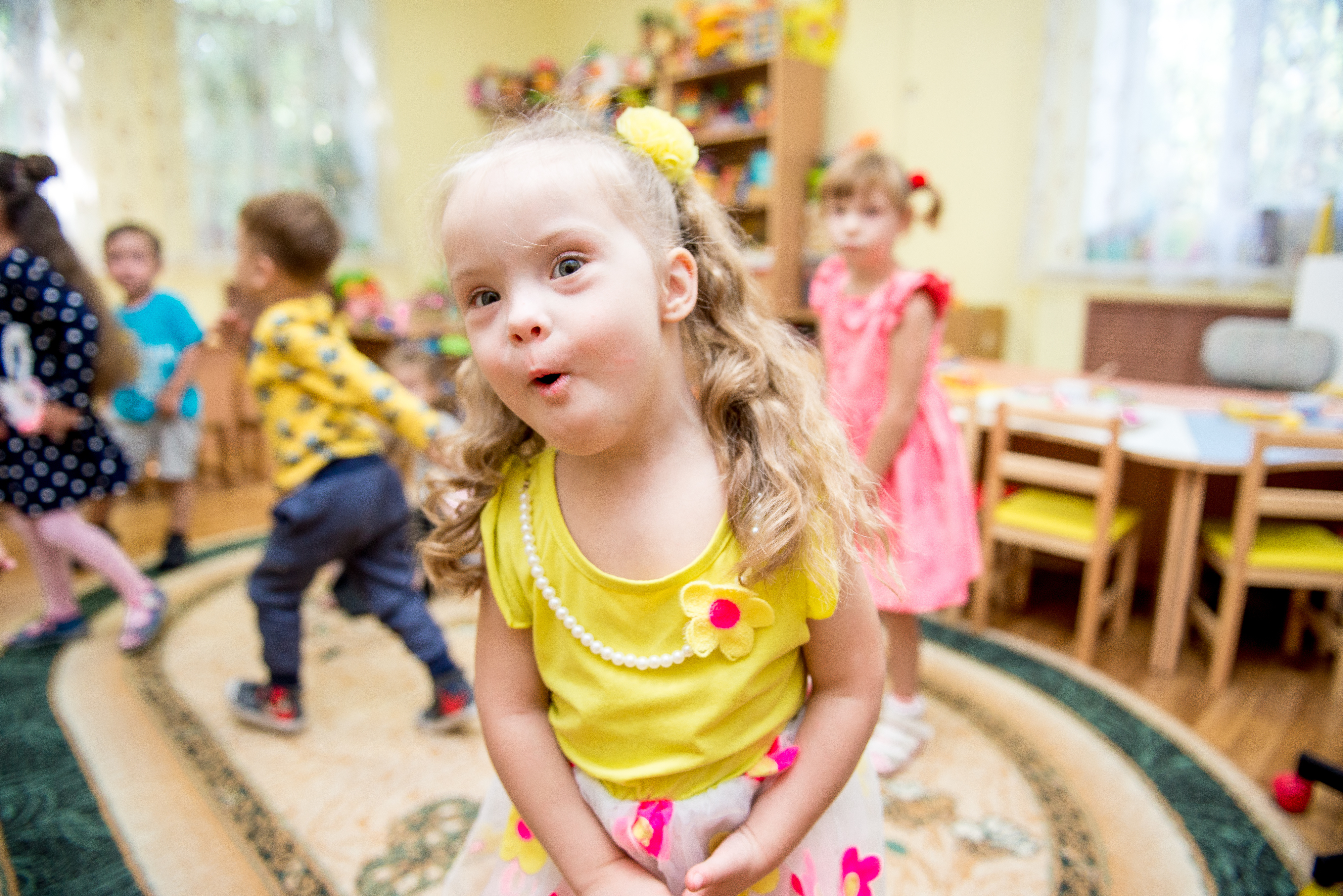 A young girl with blonde hair is playing at a preschool. She is wearing a yellow singlet top, a pearl necklace and a yellow scrunchie in her hair. There are other children wearing colourful clothes in the background.