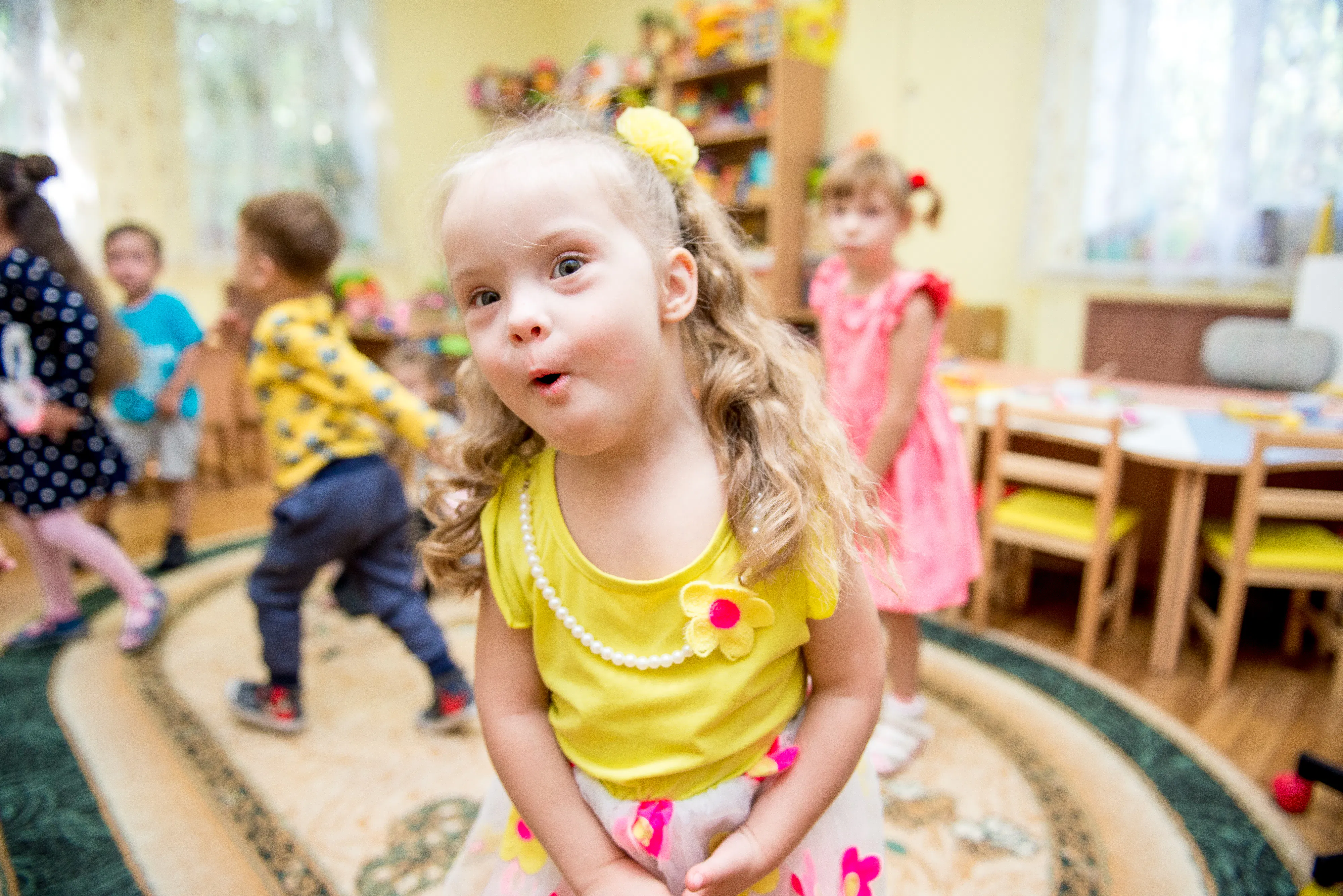 A young girl with blonde hair is playing at a preschool. She is wearing a yellow singlet top, a pearl necklace and a yellow scrunchie in her hair. There are other children wearing colourful clothes in the background.