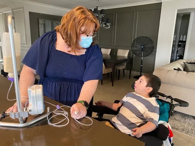 Domenico and his support worker Alax are getting ready to tube feed. They're looking at each other, and Alax is standing up, Domenico is sitting in his wheelchair. 