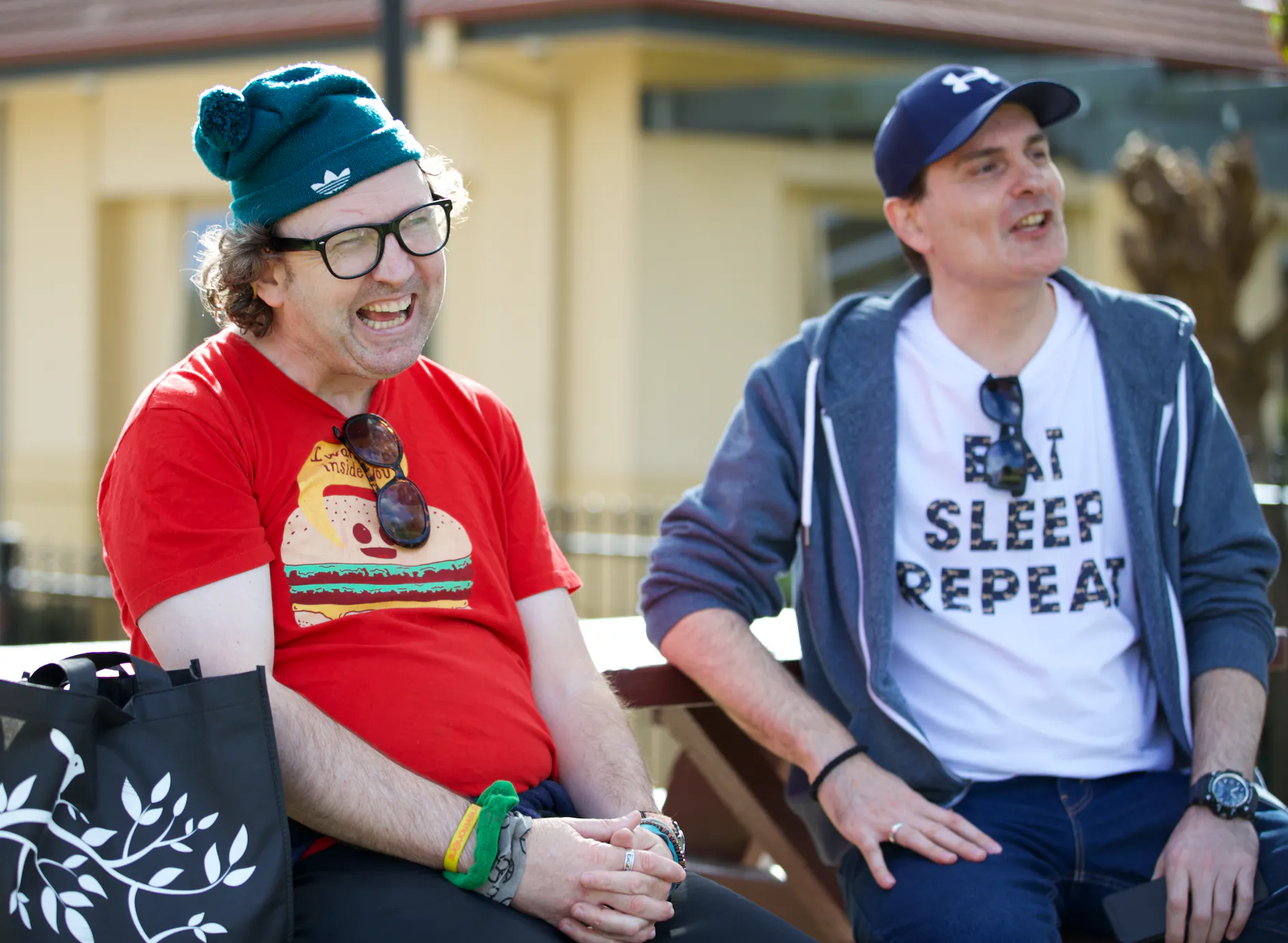 David and Goran are sitting together on a bench smiling. David is wearing a green beanie, red t-shirt and glasses. Goran is wearing a blue cap, a white t-shirt and blue hoodie.
