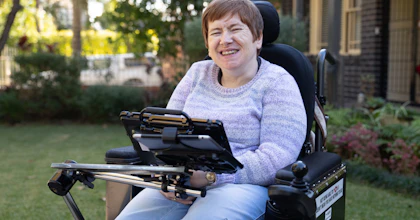 Fiona is wearing a purple knitted jumper and is smiling. She is pictured in a garden at the front of a brick building. Fiona is a wheelchair user and has an alternative and augmentative communication device.