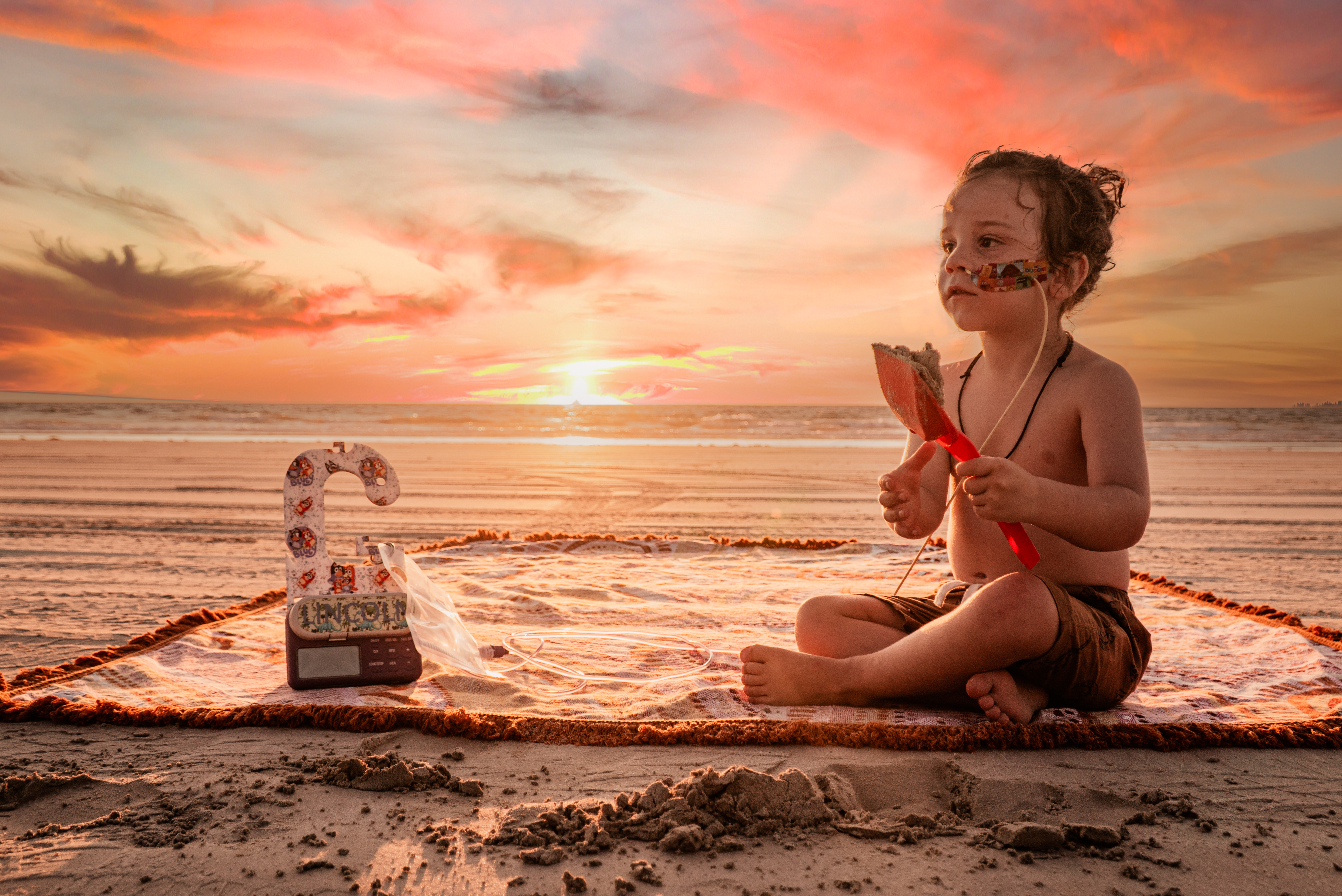 A young boy with an NG tube and tube-feeding pump sits on a beach in Western Australia as the sun sets. 