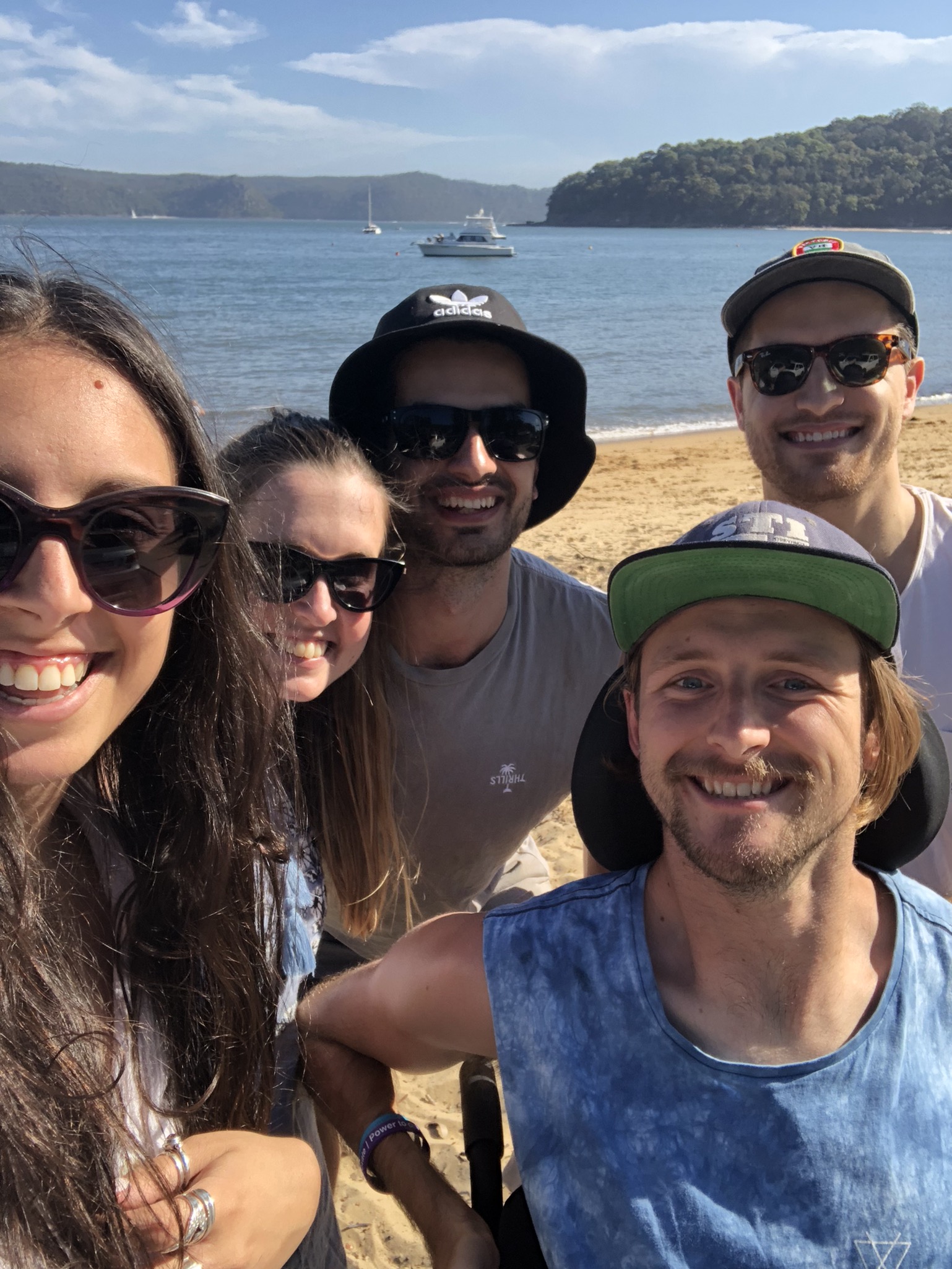 Steve is on the shore of the beach with four of his friends. He is wearing a dark cap and everyone is smiling.