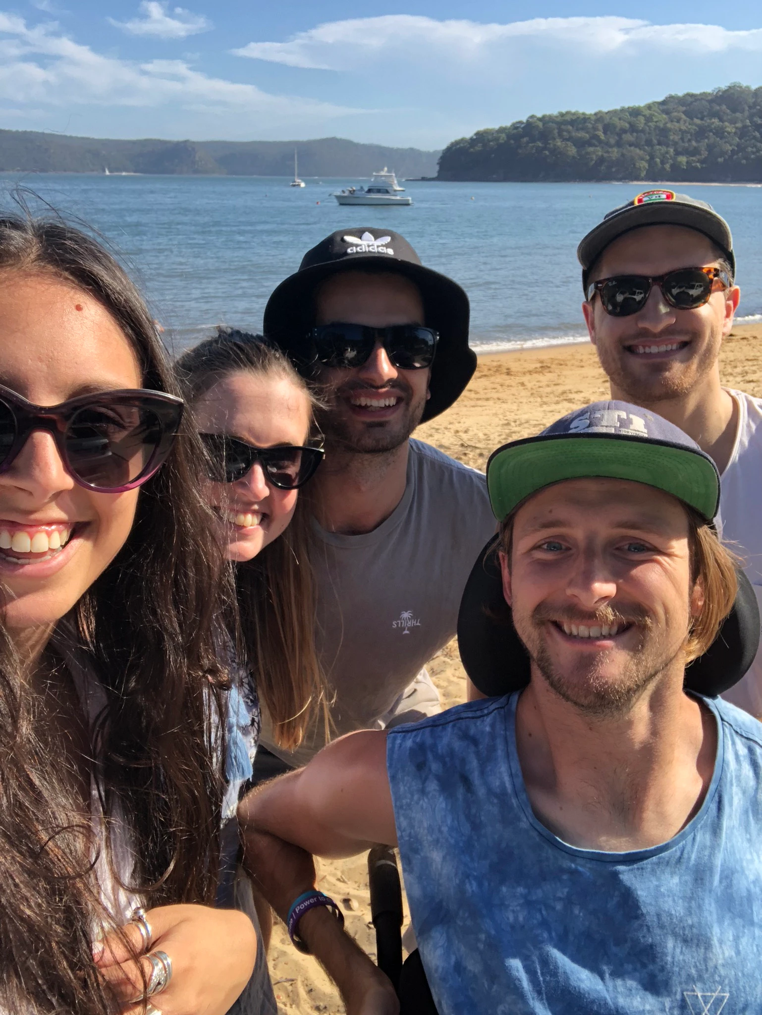Steve is on the shore of the beach with four of his friends. He is wearing a dark cap and everyone is smiling.