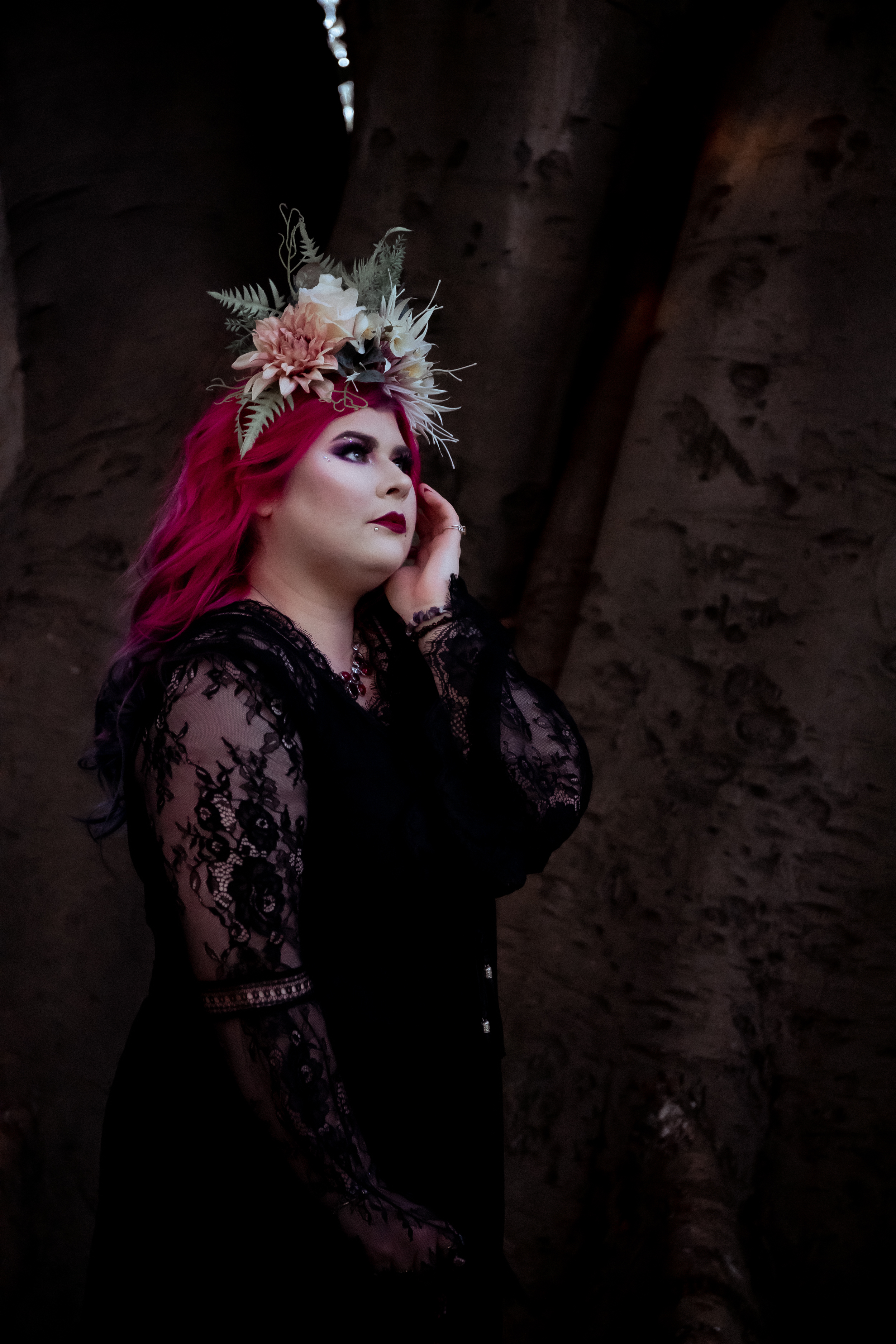 Disability advocate Zoe Simmons has long pink hair and wears a flower crown and black lace dress. She stands in front of a tree trunk and holds one hand against her face. Photo by Emma Veness