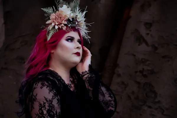 Disability advocate Zoe Simmons has long pink hair and wears a flower crown and black lace dress. She stands in front of a tree trunk and holds one hand against her face. Photo by Emma Veness