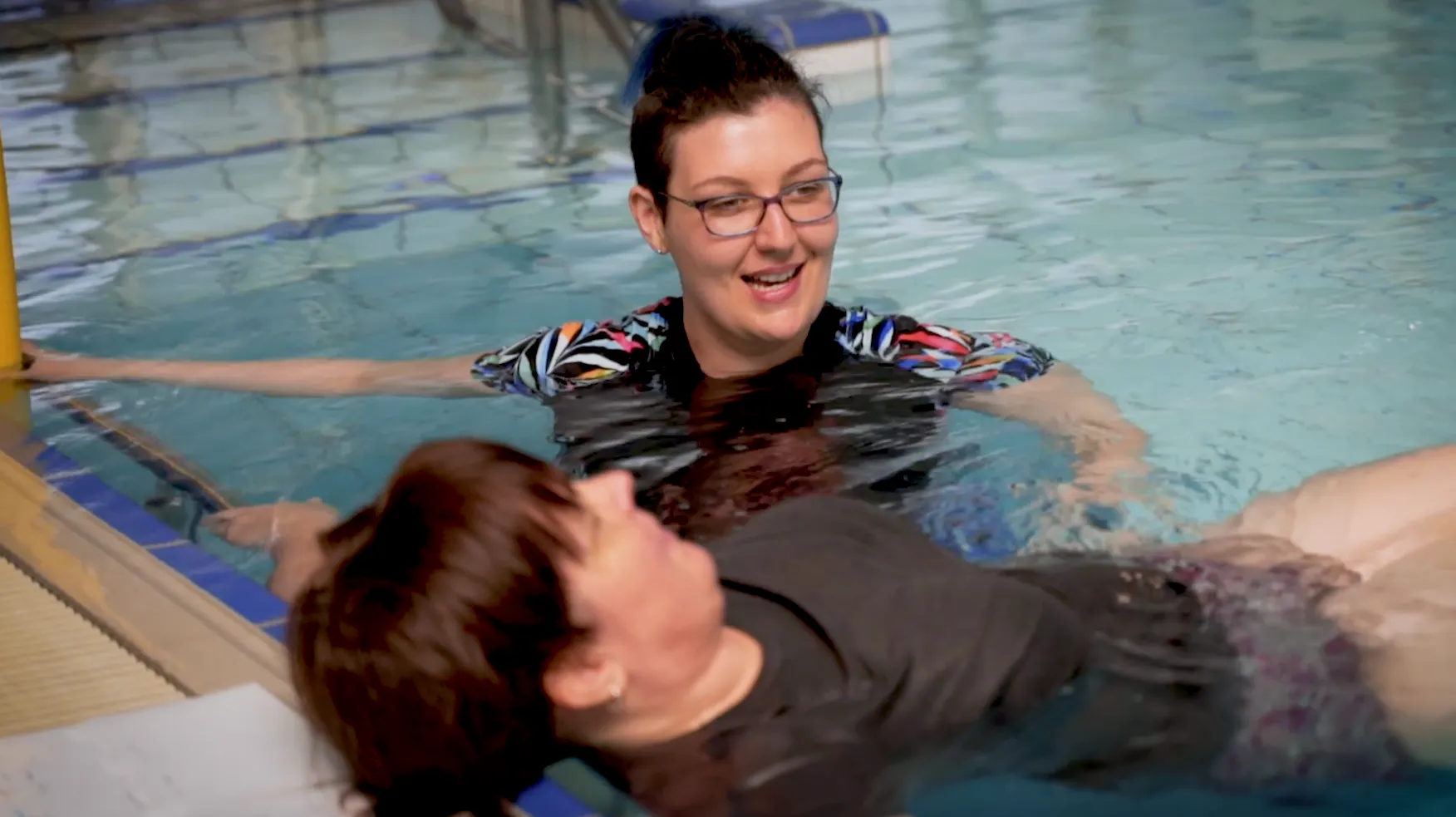 Katie and Janelle are in the pool together. Katie is assisting Janelle with her exercises.