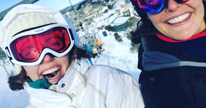 Sam is on a ski lift next to her Hireup support worker. They are both wearing ski goggles.