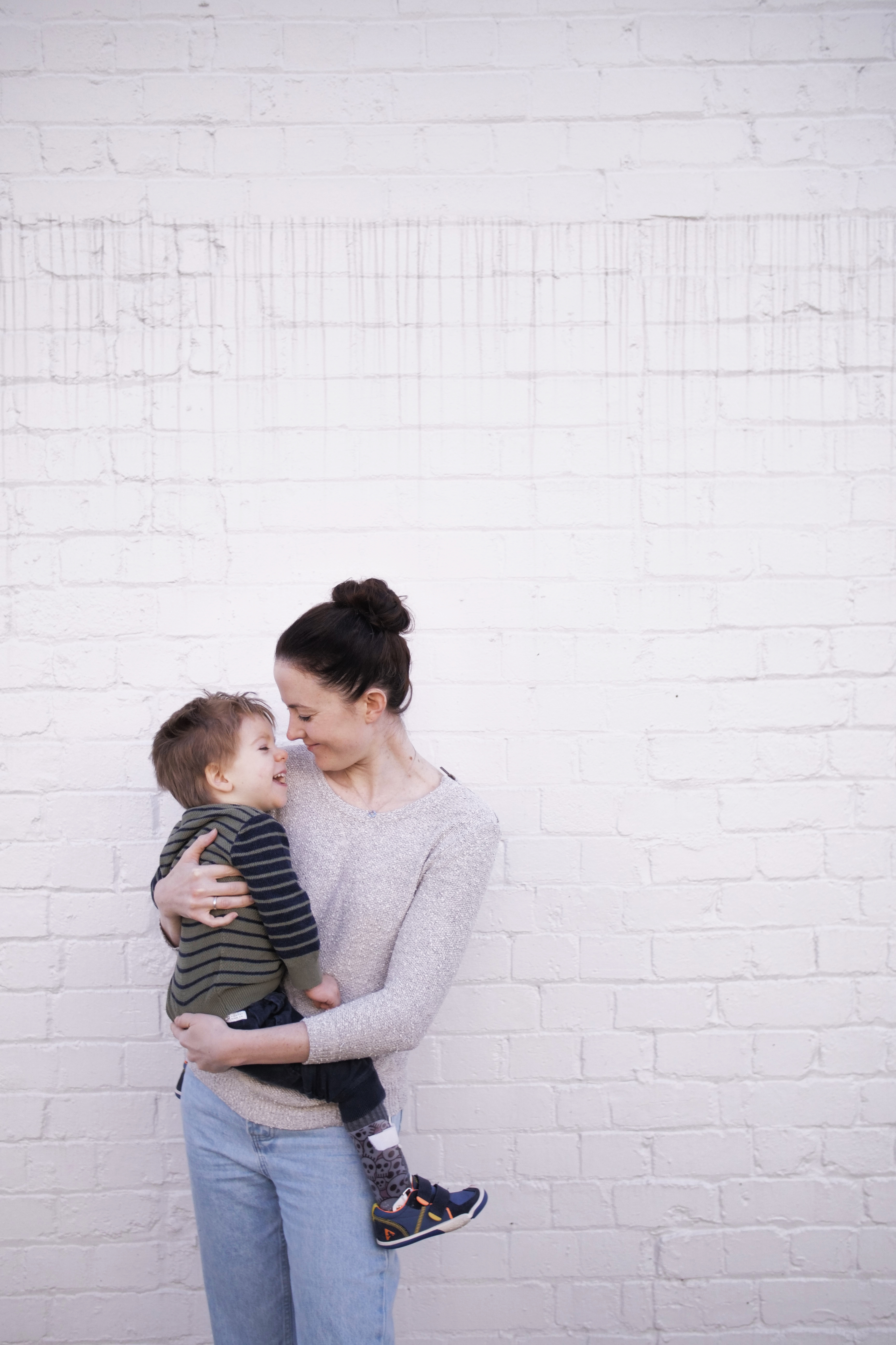 Mel, a woman with dark hair is holding her son. Her son Arlo is wearing a striped jumper and joggers. Both of them are laughing. 
