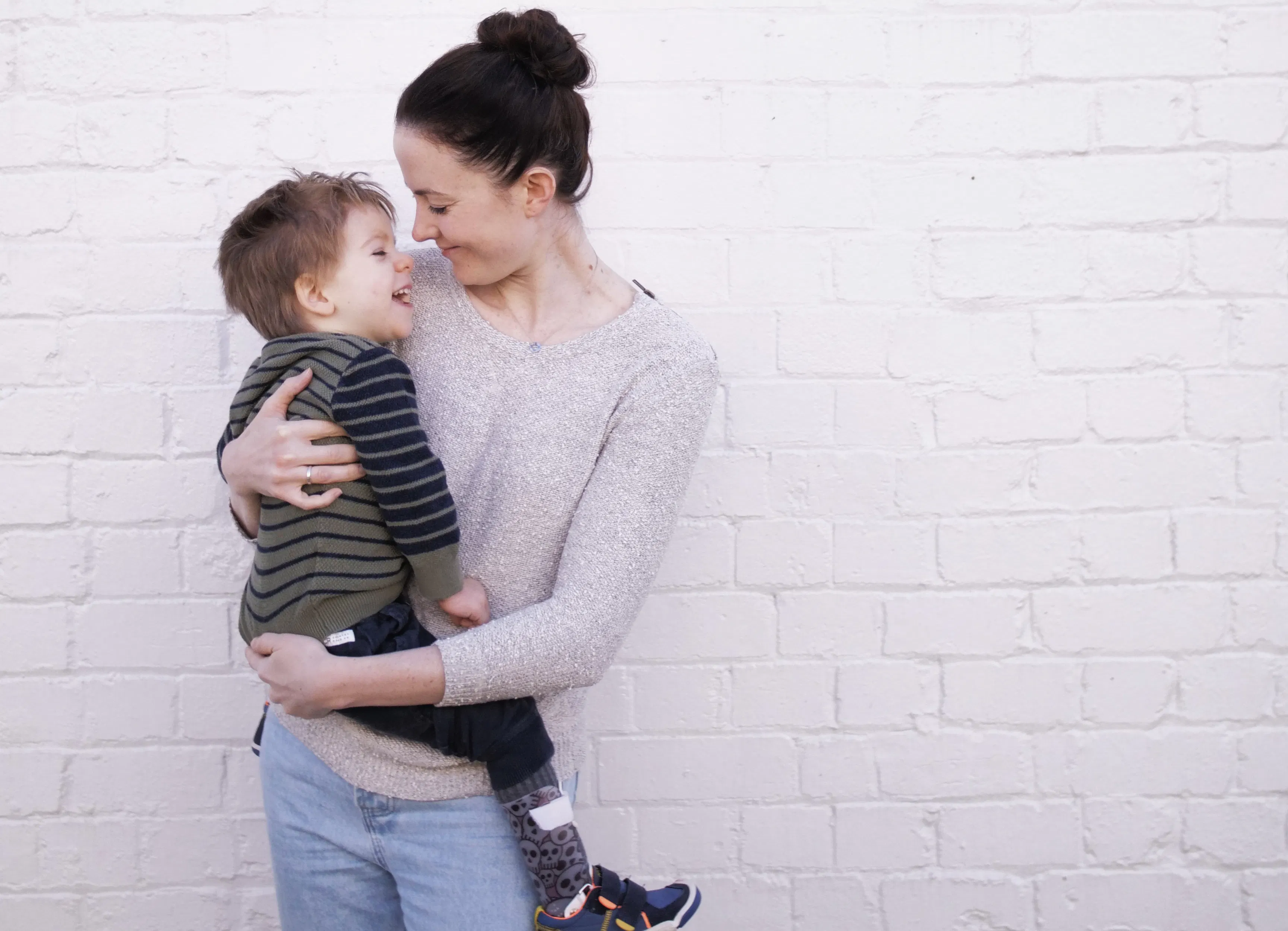 Mel, a woman with dark hair is holding her son. Her son Arlo is wearing a striped jumper and joggers. Both of them are laughing.