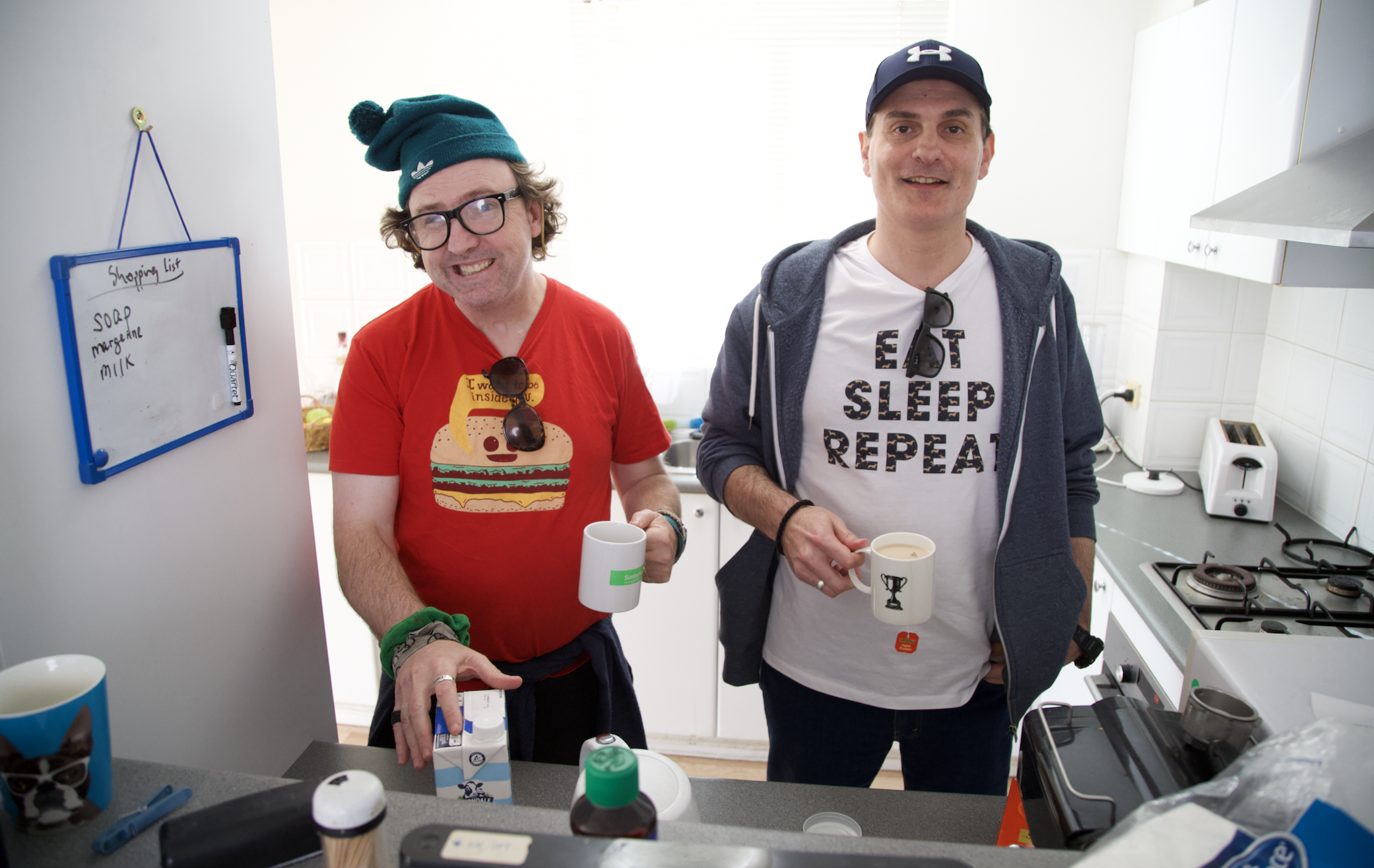 David and Goran are standing together in the kitchen. They are both smiling and holding mugs. David is reaching for a milk carton and is wearing a red t-shirt with a burger print, a green beanie and reading glasses. Goran is wearing a  blue cap, blue hoodie and white t-shirt that says eat sleep repeat. 