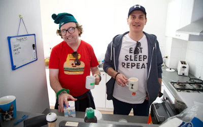 David and Goran are standing together in the kitchen. They are both smiling and holding mugs. David is reaching for a milk carton and is wearing a red t-shirt with a burger print, a green beanie and reading glasses. Goran is wearing a blue cap, blue hoodie and white t-shirt that says eat sleep repeat.