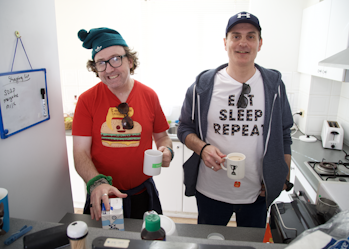 David and Goran are standing together in the kitchen. They are both smiling and holding mugs. David is reaching for a milk carton and is wearing a red t-shirt with a burger print, a green beanie and reading glasses. Goran is wearing a blue cap, blue hoodie and white t-shirt that says eat sleep repeat.