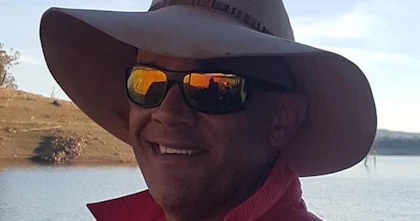 Wayne Wright is wearing a red shirt, sunglasses and hat and is smiling at the camera.