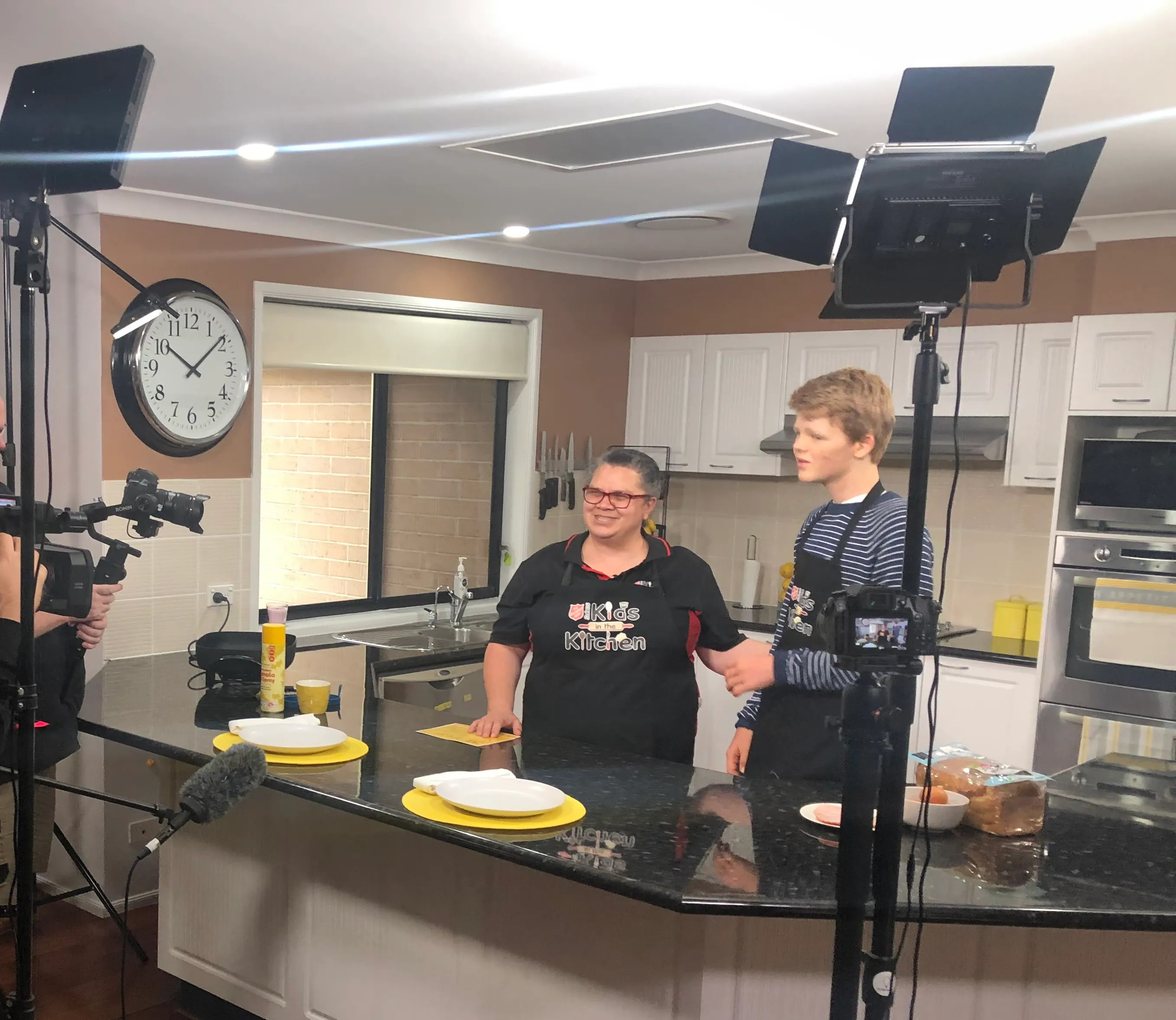 A young man and a woman are in a kitchen. They're wearing aprons and are smiling. A television crew is set up in the kitchen. 