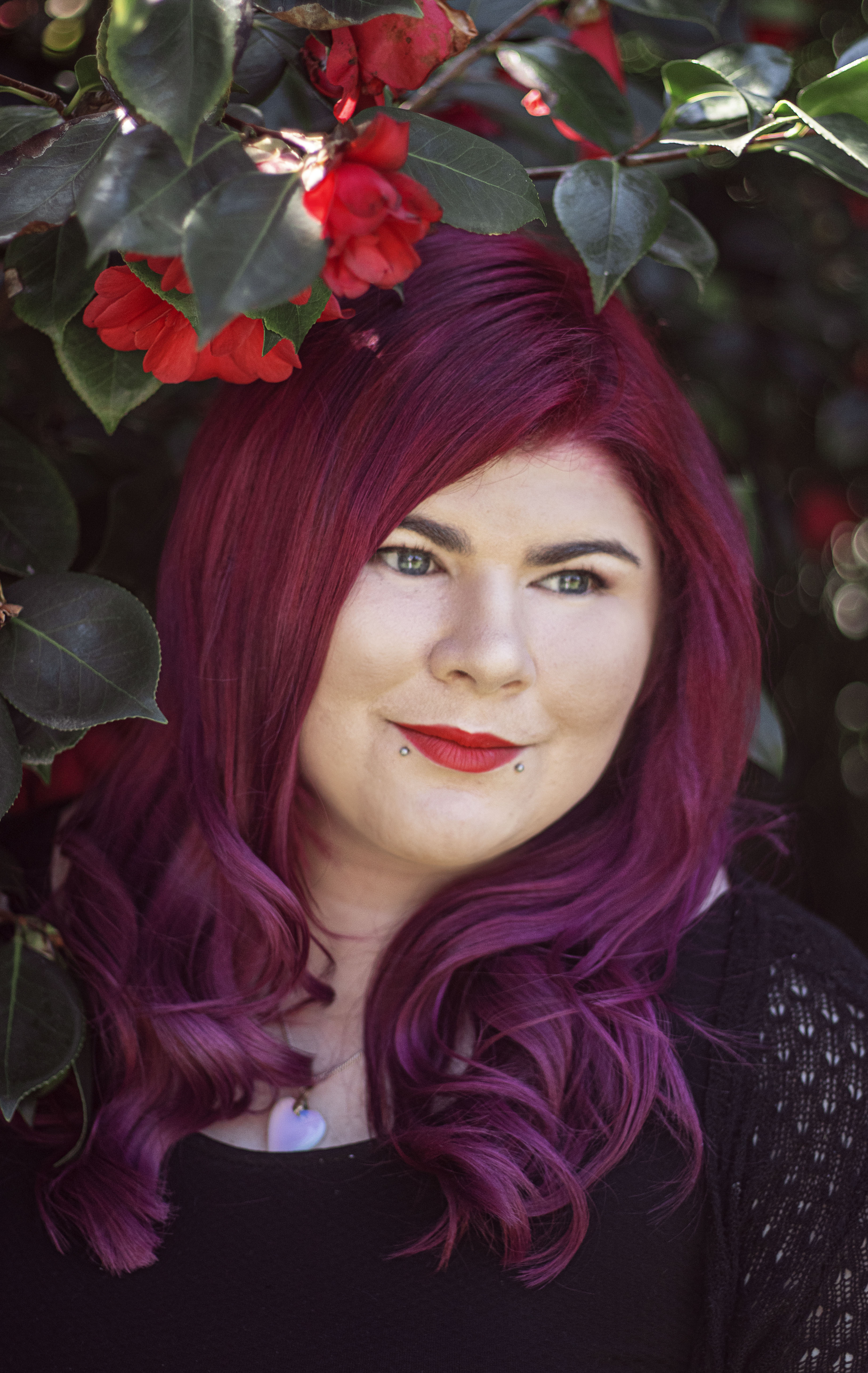 Zoe has dark red hair and is wearing red lipstick. She is standing in front of a bush with red flowers. 